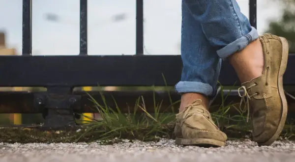 Gambe con jeans arrotolati e scarpe senza calzini, appoggiate su un muretto con erba e ghiaia.
