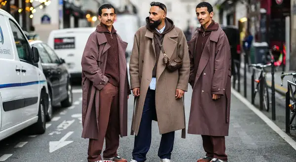 Tre uomini con eleganti stili abbigliamento uomo, cappotti lunghi e pantaloni coordinati, posano in strada.