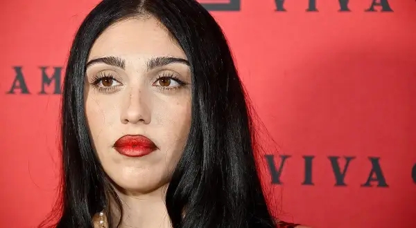 Lourdes Leon, con labbra rosse audaci e sguardo intenso, posa davanti a uno sfondo rosso con scritte nere.