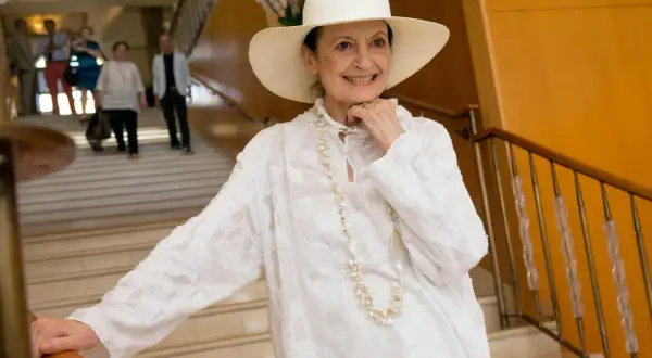 Carla Fracci sorride con un cappello bianco e una collana di perle, elegante su una scala.