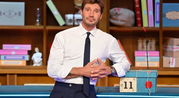 Stefano De Martino outfit: camicia bianca, cravatta blu scuro e pantaloni eleganti. Sullo sfondo, una libreria piena di oggetti colorati.