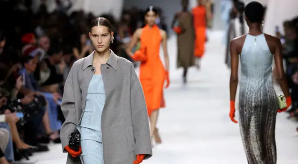 Angelina Kendall sfila con un cappotto grigio oversize e un abito a righe azzurre, guanti arancioni vivaci.