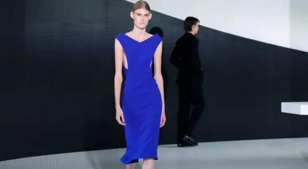Modella con abito blu elettrico, un'opera di Simone Bellotti, sfila su una passerella minimalista.