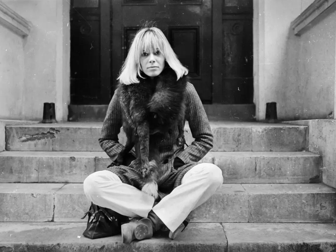 Anita Pallenberg, icona di stile, posa con un cappotto di tweed e una pelliccia voluminosa, seduta sulle scale di un portone.