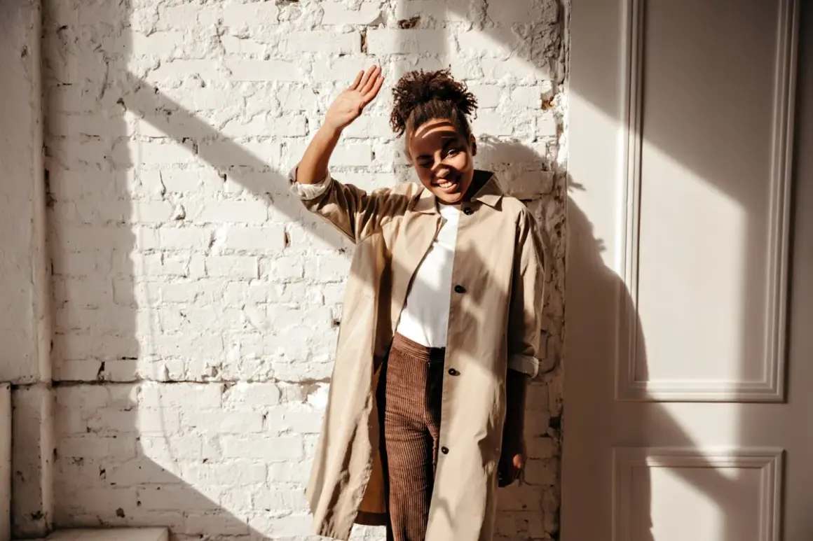 Donna con trench beige e pantaloni marroni, con la mano alzata per proteggersi dal sole. Un outfit versatile, ma quali scarpe abbinare?