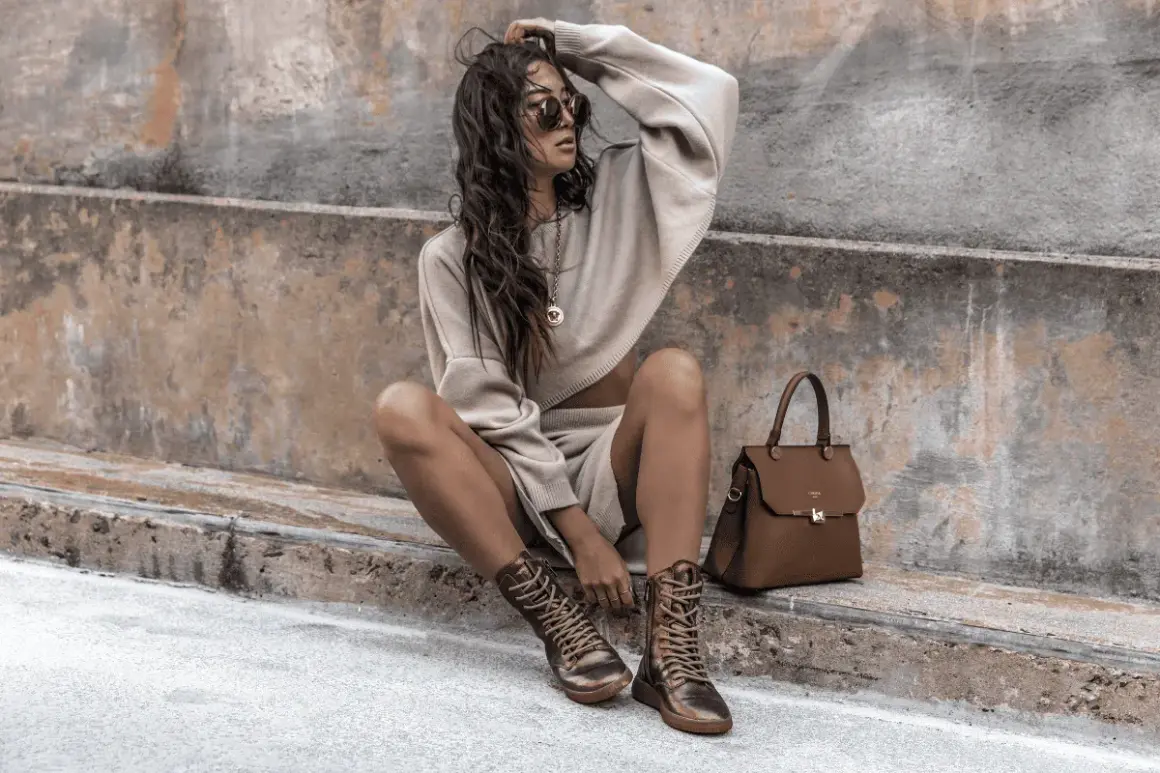 Donna con un maglione corto e pantaloncini beige, stivali stringati e borsa marrone. Sembra un uomo basso.