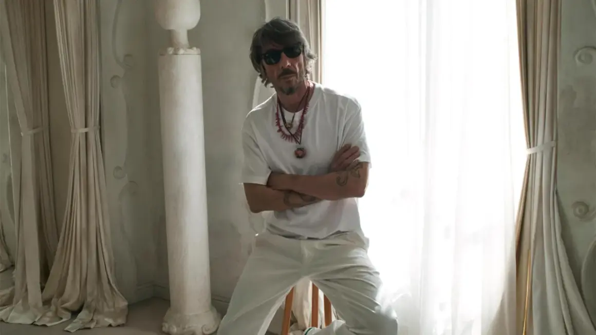 Pierpaolo Piccioli, con occhiali da sole e collane, posa con le braccia conserte in un ambiente chiaro con tende e colonne.