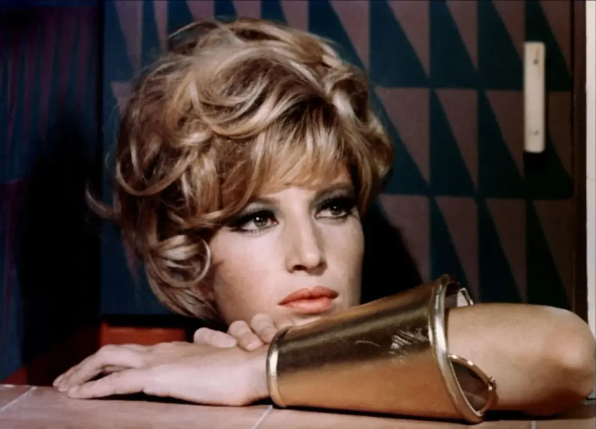Monica Vitti, con un bracciale dorato e un'espressione pensierosa, sfoggia un look audace con capelli biondi cotonati e trucco marcato.