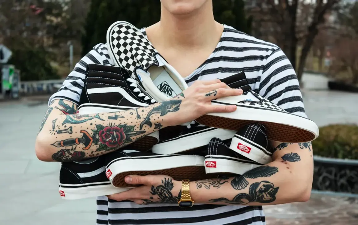 Un ragazzo con tatuaggi colorati tiene in braccio diversi tipi di Vans: Sk8-Hi, Slip-On a scacchi e altre classiche.