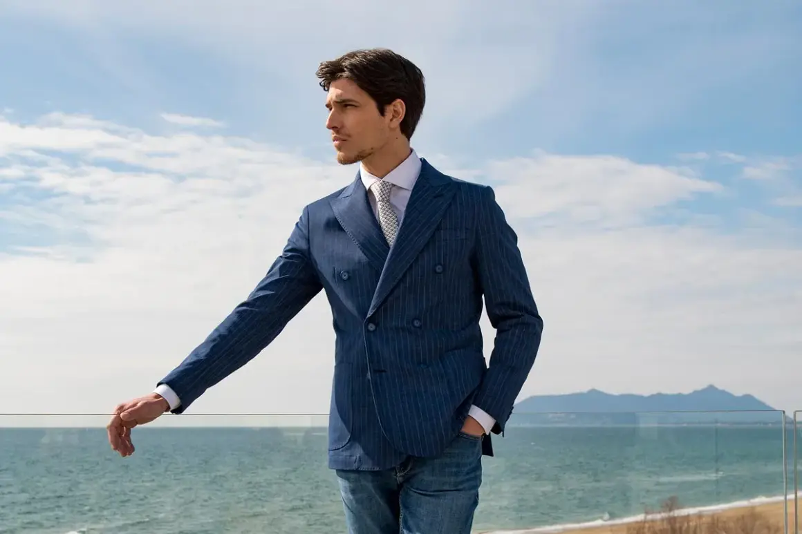Uomo con giacca blu a doppio petto a righe, camicia bianca e cravatta, appoggiato a una ringhiera con vista mare.