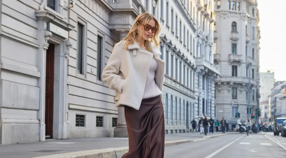 Donna con tailleur in inverno: un cappotto corto in montone bianco sopra un maglione e gonna di seta marrone.