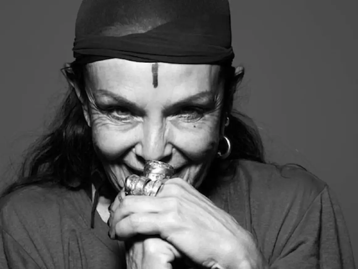Michele Lamy, con un turbante nero e un segno sulla fronte, sorride enigmaticamente, le mani adornate da anelli vistosi.