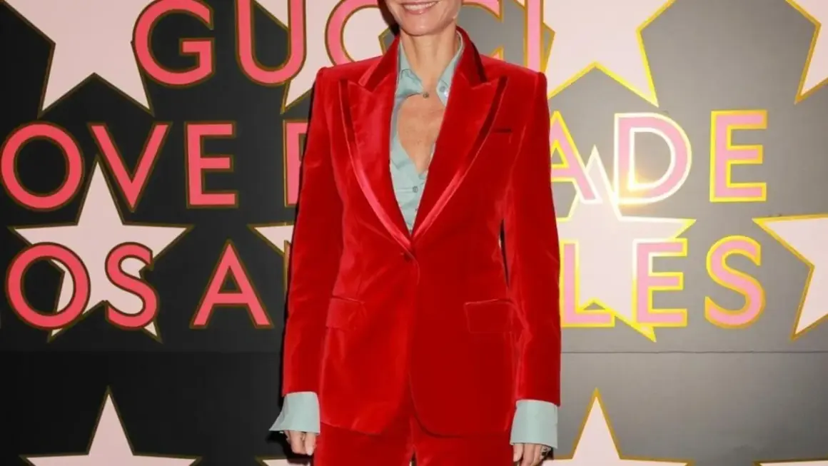 Gwyneth Paltrow in un completo rosso di velluto, sorride davanti a uno sfondo con stelle e lettere rosa e oro.