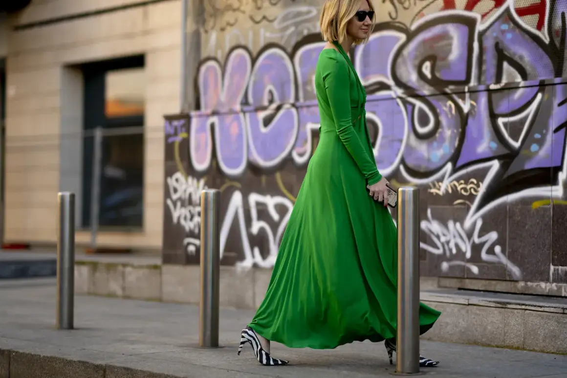Donna con vestito lungo elegante verde smeraldo e scarpe zebrate, cammina con passo deciso davanti a un muro con graffiti.