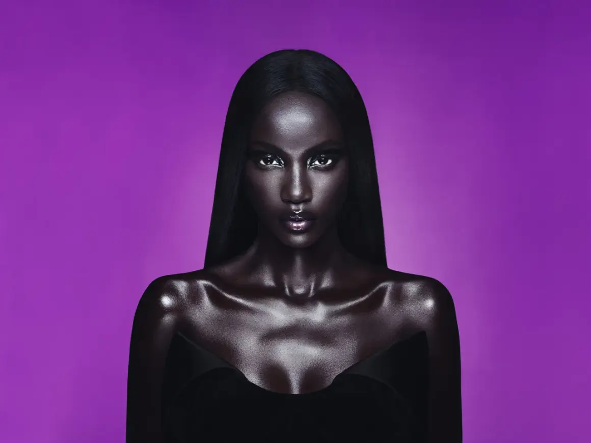 Anok Yai, con pelle lucida e capelli neri, posa con uno sguardo intenso su sfondo viola.