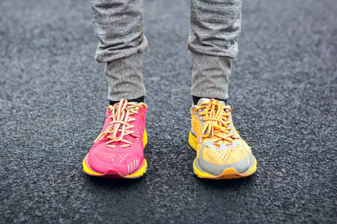 Due diversi tipi di scarpe da ginnastica, una rosa acceso e una arancione, indossate con pantaloni grigi su una superficie scura.