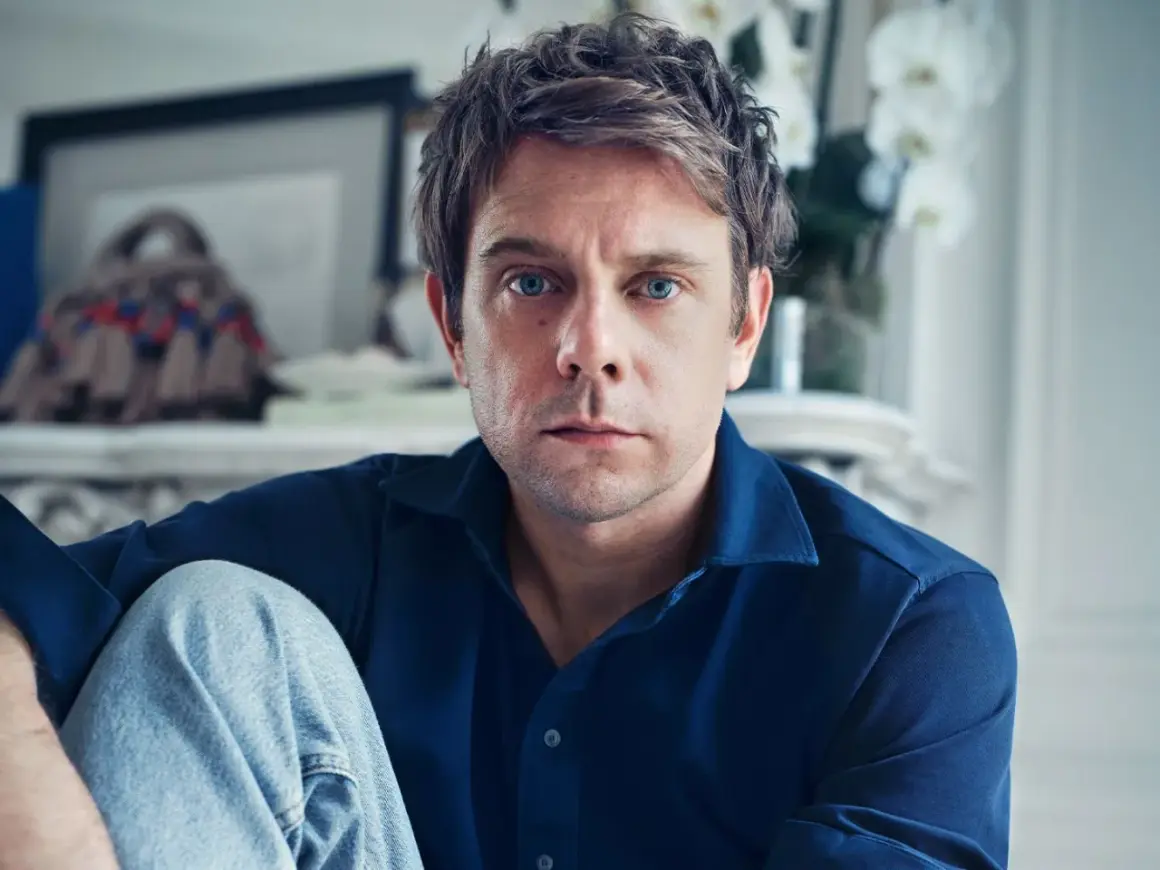 Ritratto di Jonathan Anderson, con occhi azzurri penetranti, indossa una camicia blu e jeans.