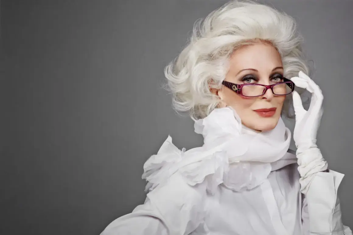 Carmen Dell'Orefice, icona di stile, con capelli bianchi voluminosi, occhiali viola e guanti bianchi, posa con eleganza.