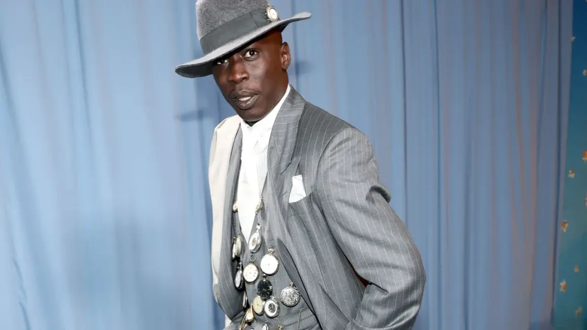 Un uomo con un cappello grigio e un completo gessato, ornato da numerosi orologi da taschino, incarna l'eleganza maschile.