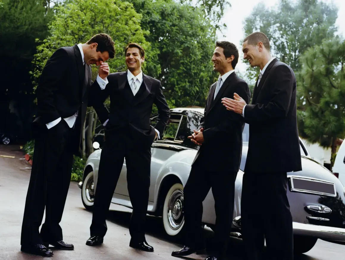 Quattro uomini in abito scuro, perfetti per un matrimonio, ridono vicino a un'auto d'epoca.