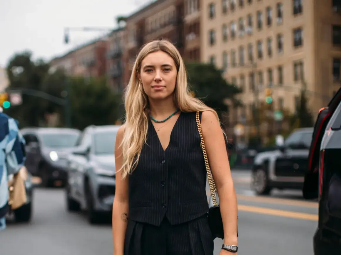 Donna con capelli biondi indossa un elegante gilet doppiopetto a righe, collana turchese e borsa a tracolla dorata, in strada.