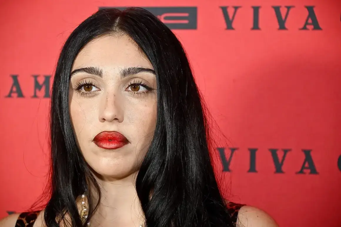 Lourdes Leon, con labbra rosse audaci e sguardo intenso, posa davanti a uno sfondo rosso con scritte nere.
