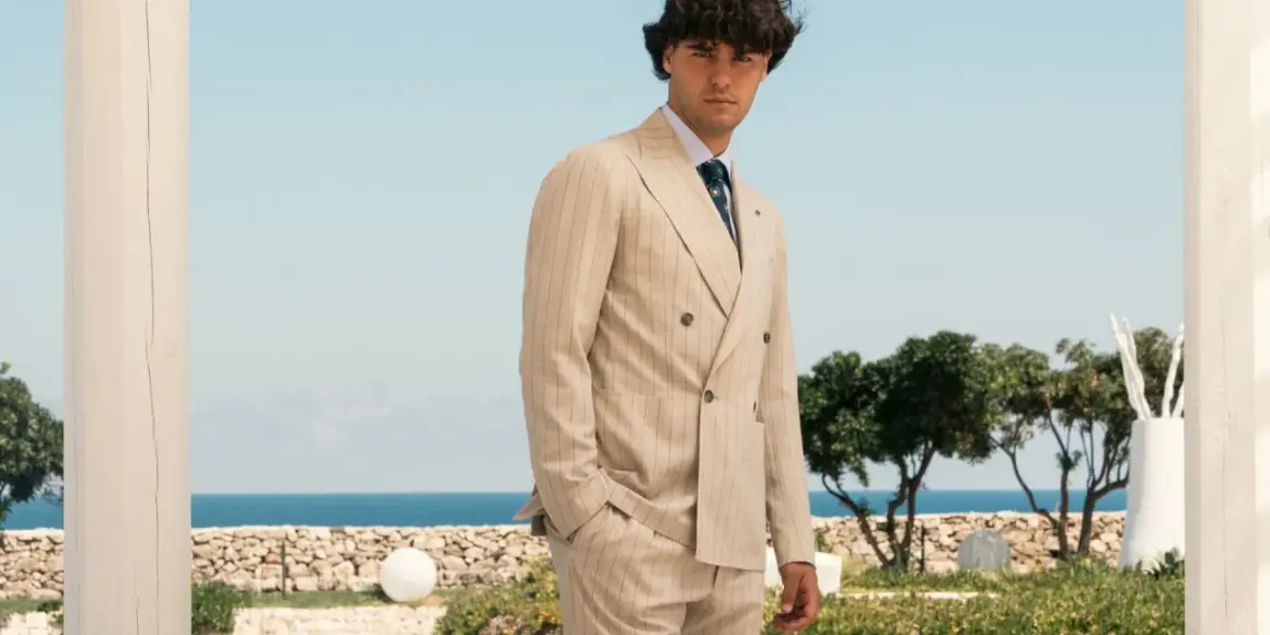 Uomo elegante con abito gessato beige, in piedi con lo sguardo rivolto verso l'osservatore, con il mare e il cielo azzurro sullo sfondo.