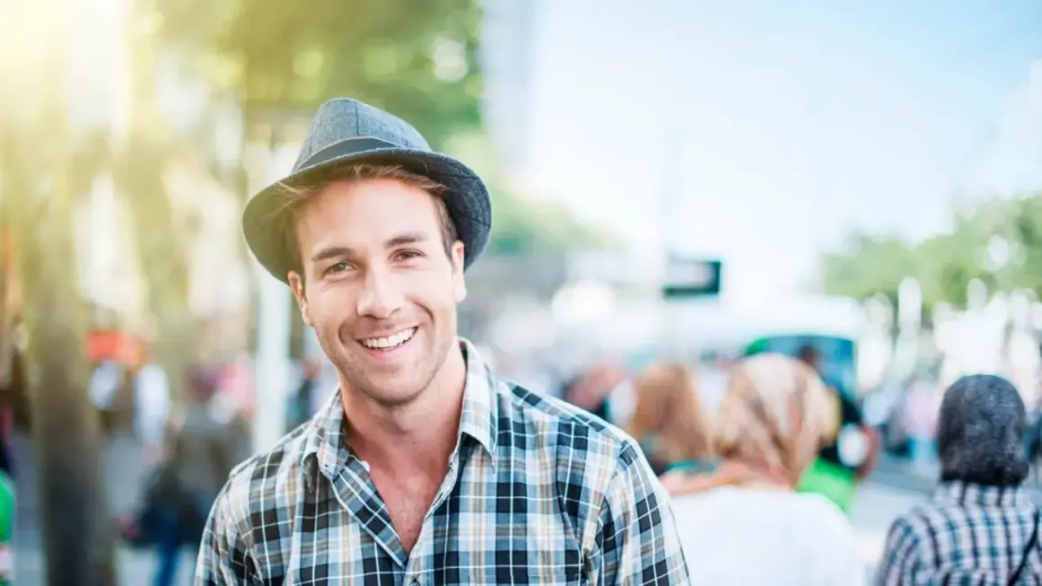 Uomo sorridente con un cappello adatto al viso, indossa una camicia a quadri. La luce del sole illumina la scena.