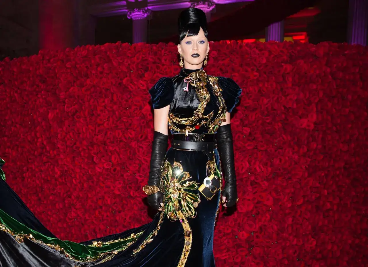 Katy Perry al Met Gala, con un abito scuro e dettagli dorati, posa davanti a uno sfondo di rose rosse.