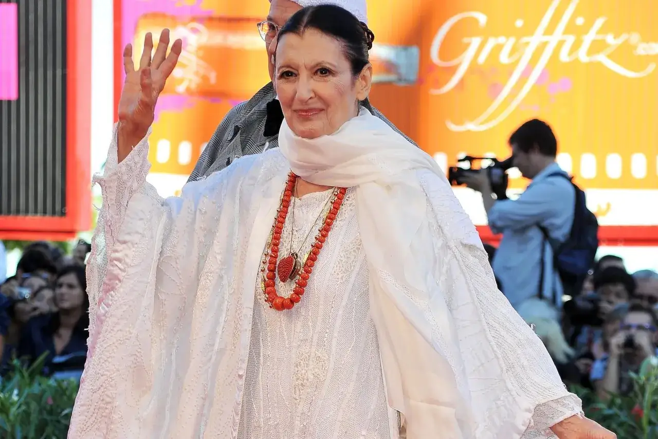 Carla Fracci saluta con la mano, radiosa in abito bianco e collana di corallo.