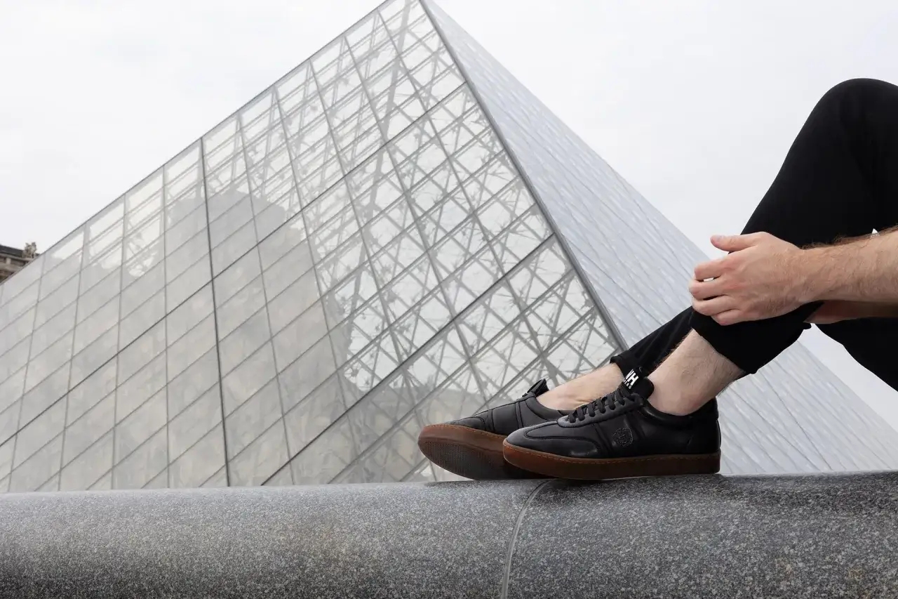 Un uomo indossa un outfit con sneakers nere, seduto davanti alla piramide del Louvre.