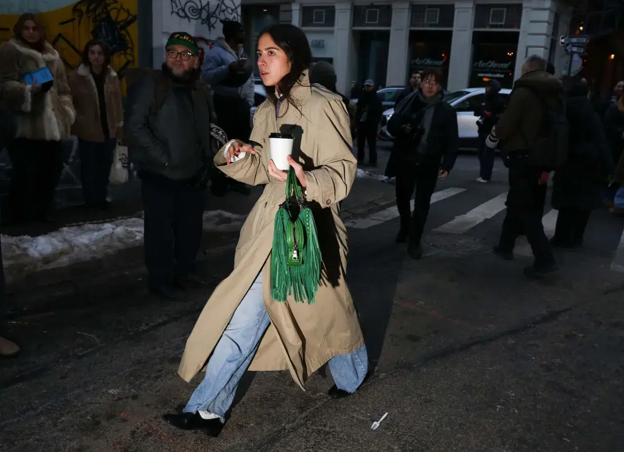 Una donna cammina per strada con un caff&egrave; in mano, indossando un classico trench beige, jeans e una borsa verde con frange.