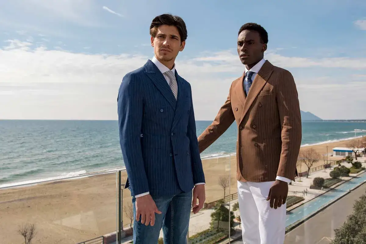 Due uomini con eleganti stili abbigliamento uomo, uno in blu gessato, l'altro in marrone, posano con il mare sullo sfondo.