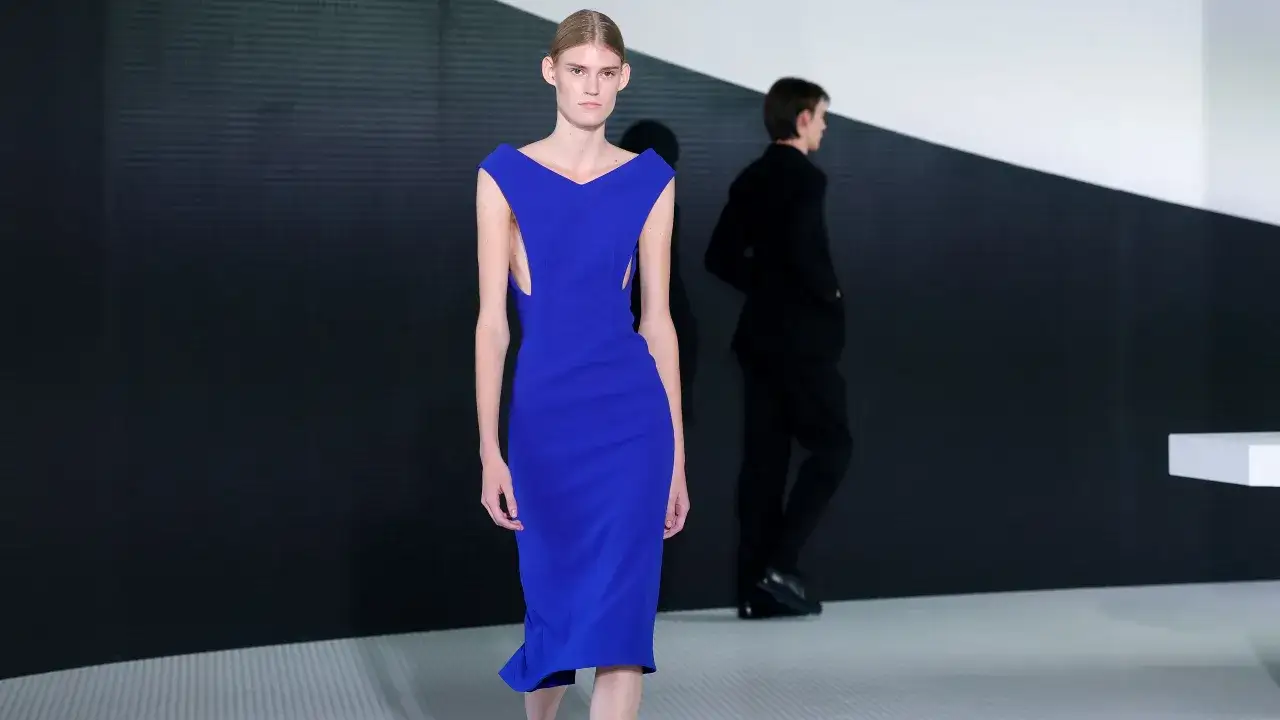 Modella con abito blu elettrico su passerella, con un uomo in abito nero sullo sfondo. Simone Bellotti cattura l'eleganza.