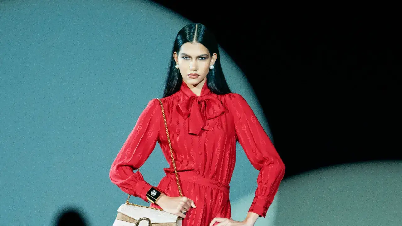 Modella con abito rosso Gucci, borsa a tracolla e gioielli vistosi, pronta per la sfilata.