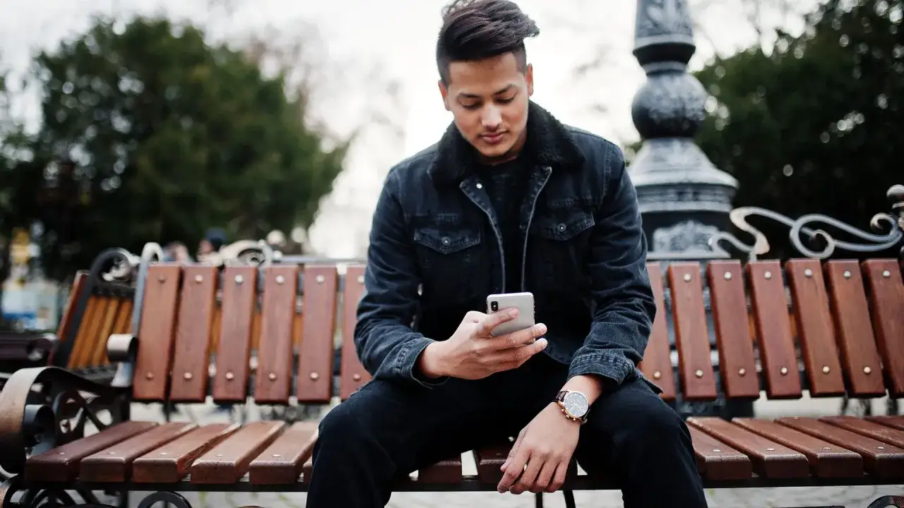 Giovane uomo con abbinamento jeans nero uomo, giacca di jeans scura e orologio, seduto su una panchina di legno all'aperto, guarda il suo smartphone.