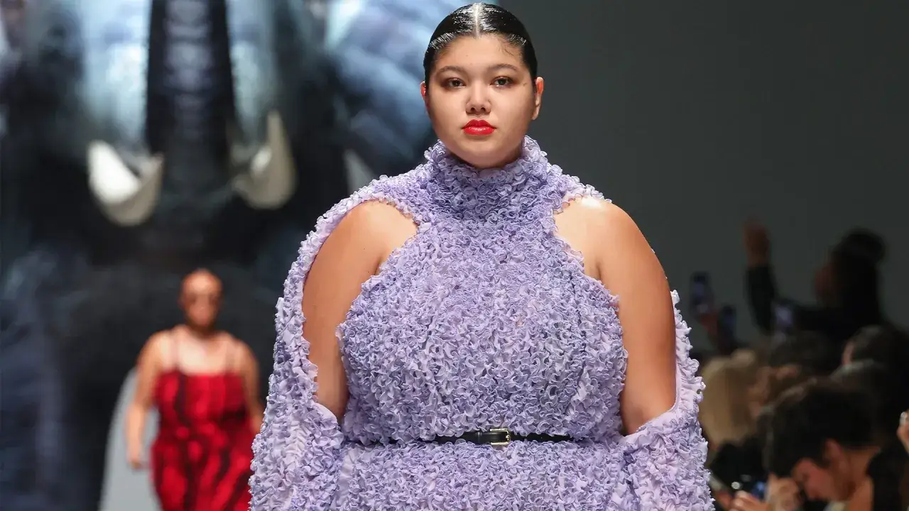 Modella curvy sfila con un abito lilla voluminoso e dettagliato. Sullo sfondo, un'altra modella curvy in rosso.
