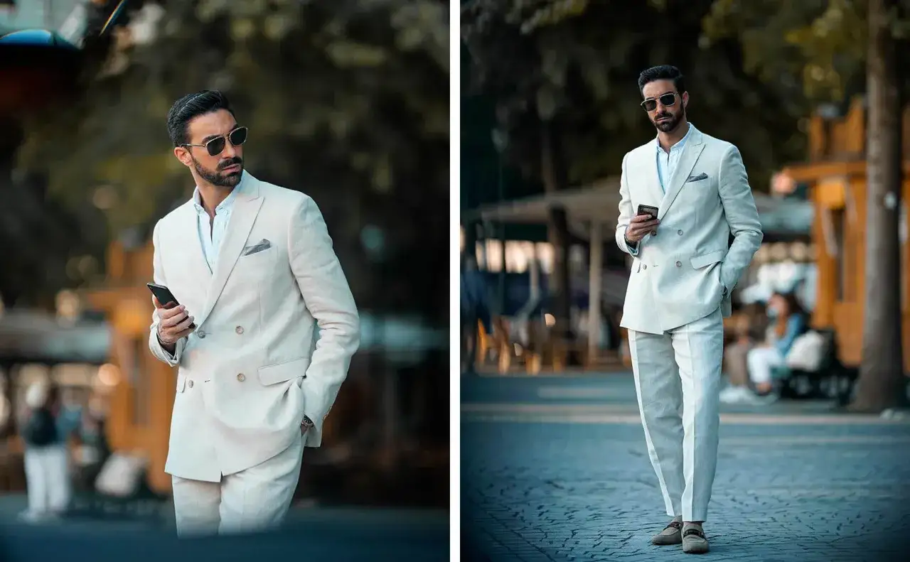 Uomo con abito semi formal color crema, occhiali da sole e smartphone in mano, passeggia in una strada acciottolata.