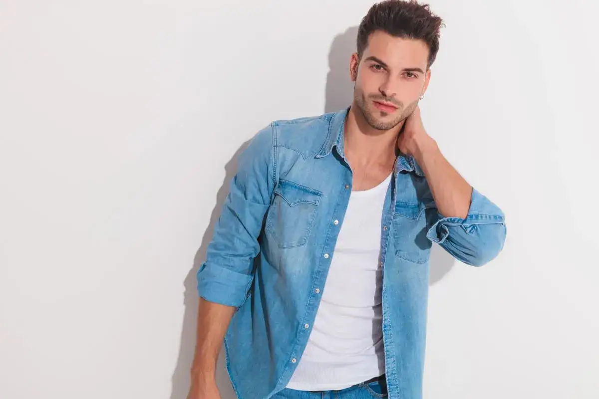 Uomo con jeans e camicia di jeans aperta su una canottiera bianca, con una mano dietro il collo.