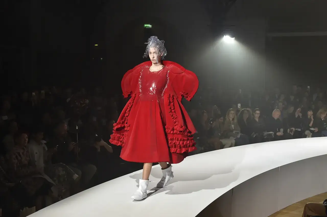 Modella con un abito rosso voluminoso e futuristico, un'opera d'arte di Rei Kawakubo, sfila su una passerella illuminata.