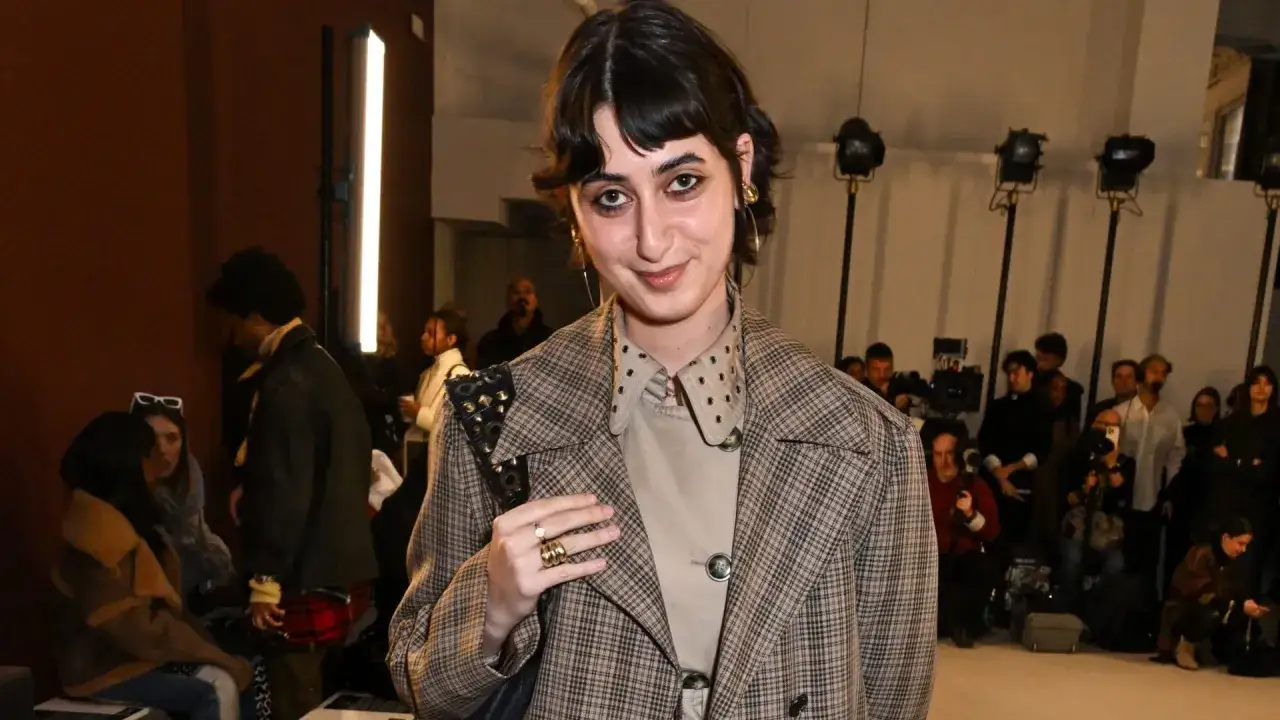 Giovane donna con un cappotto a quadri, un look che ricorda rama duwaji, sorride tra la folla di fotografi.