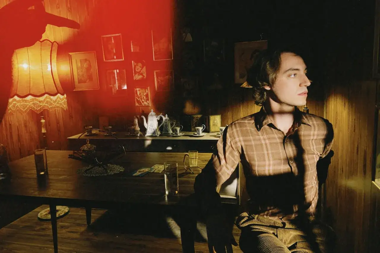 Luca Vergoni, in un interno con lampada rossa e foto alle pareti, guarda pensieroso fuori campo.