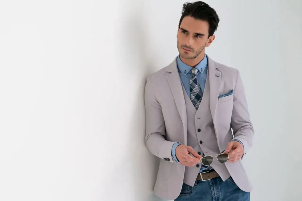 Uomo elegante con invitato abito cerimonia estivo: completo grigio chiaro, camicia azzurra, cravatta a quadri e jeans.