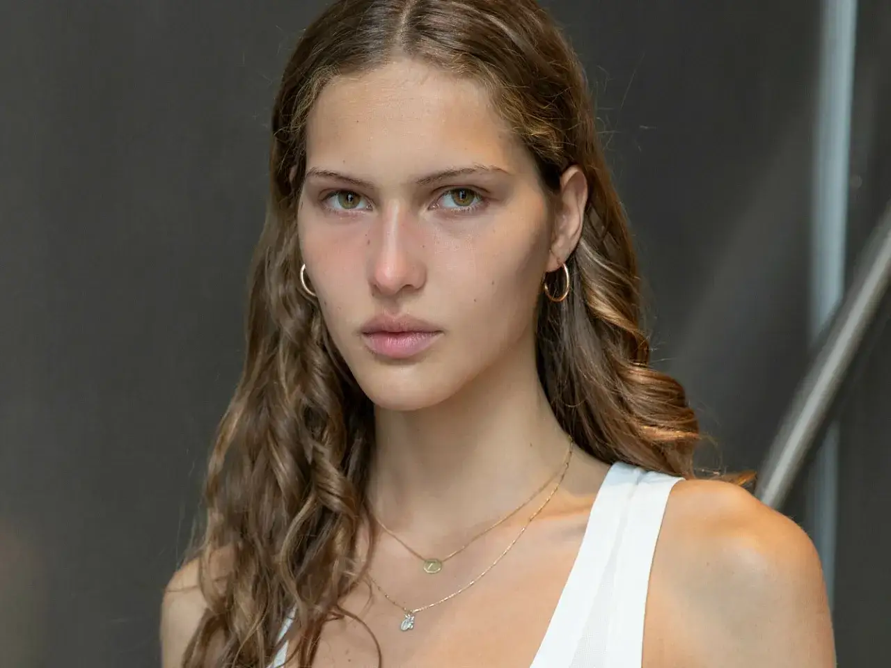 Ritratto di Angelina Kendall con capelli castani mossi, orecchini a cerchio e collane dorate.