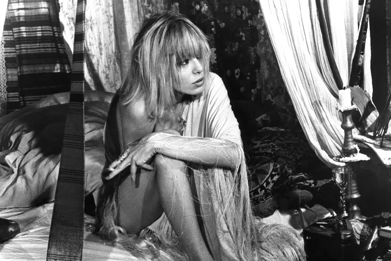 Anita Pallenberg, icona di stile e musa rock, posa in un letto dall'atmosfera bohémien, circondata da tessuti e candele.
