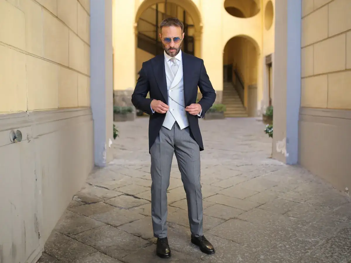 Uomo elegante in spezzato estivo per matrimonio, con giacca blu, gilet e pantaloni grigi, in un cortile con archi.