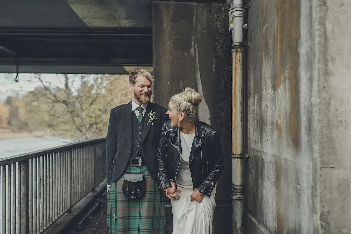 Rebecca Donaldson e il suo sposo, con kilt scozzese, ridono sotto un ponte.