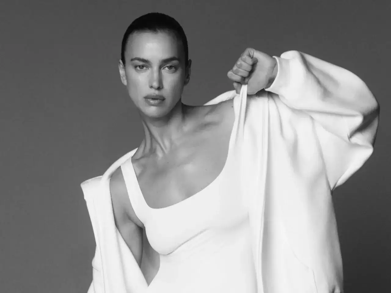 Irina Shayk, con un top bianco e una felpa aperta, posa con un'espressione intensa.