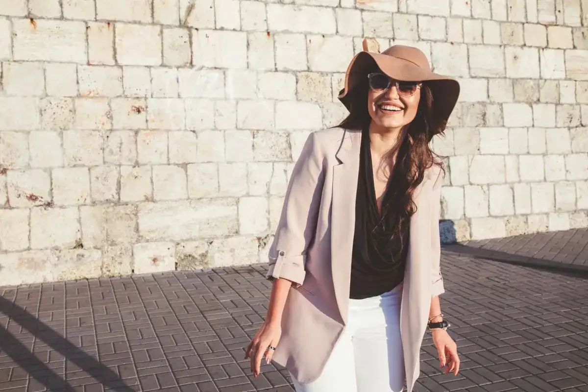 Donna sorridente con cappello e occhiali da sole, perfetta per come vestirsi con 20 gradi: blazer leggero, top nero e pantaloni bianchi.