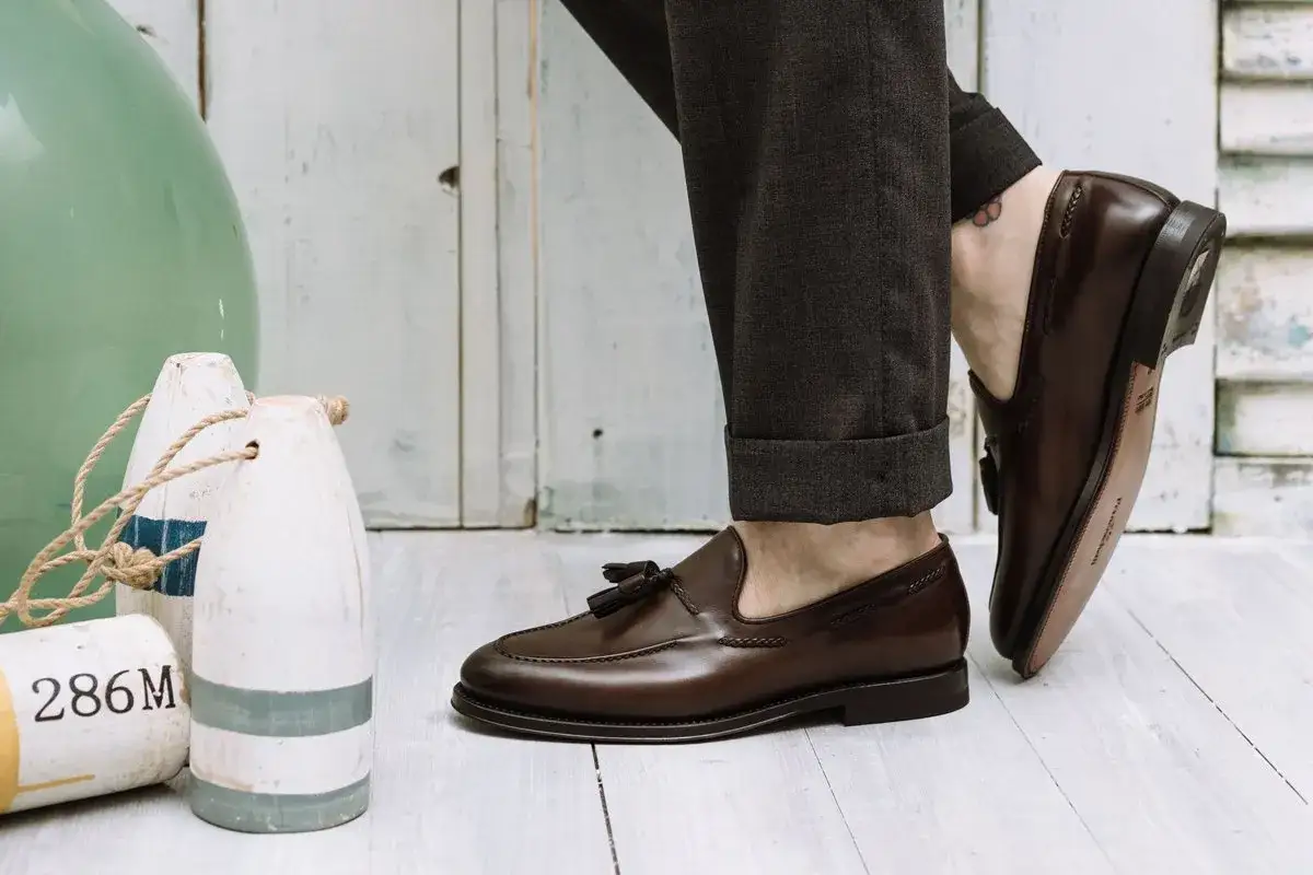 Un uomo elegante indossa mocassini marroni con nappine, abbinati a pantaloni scuri arrotolati.